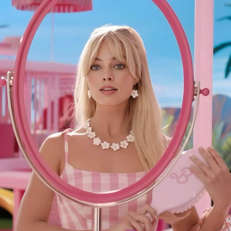 A picture of Margot Robbie as Barbie in a gingham dress and floral jewellery, brushing her hair into a Barbie mirror.